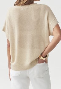 Top a maniche corte lavorato a maglia beige indossato con pantaloni bianchi, bracciale dorato sul polso, modello visto di spalle con una mano in tasca.