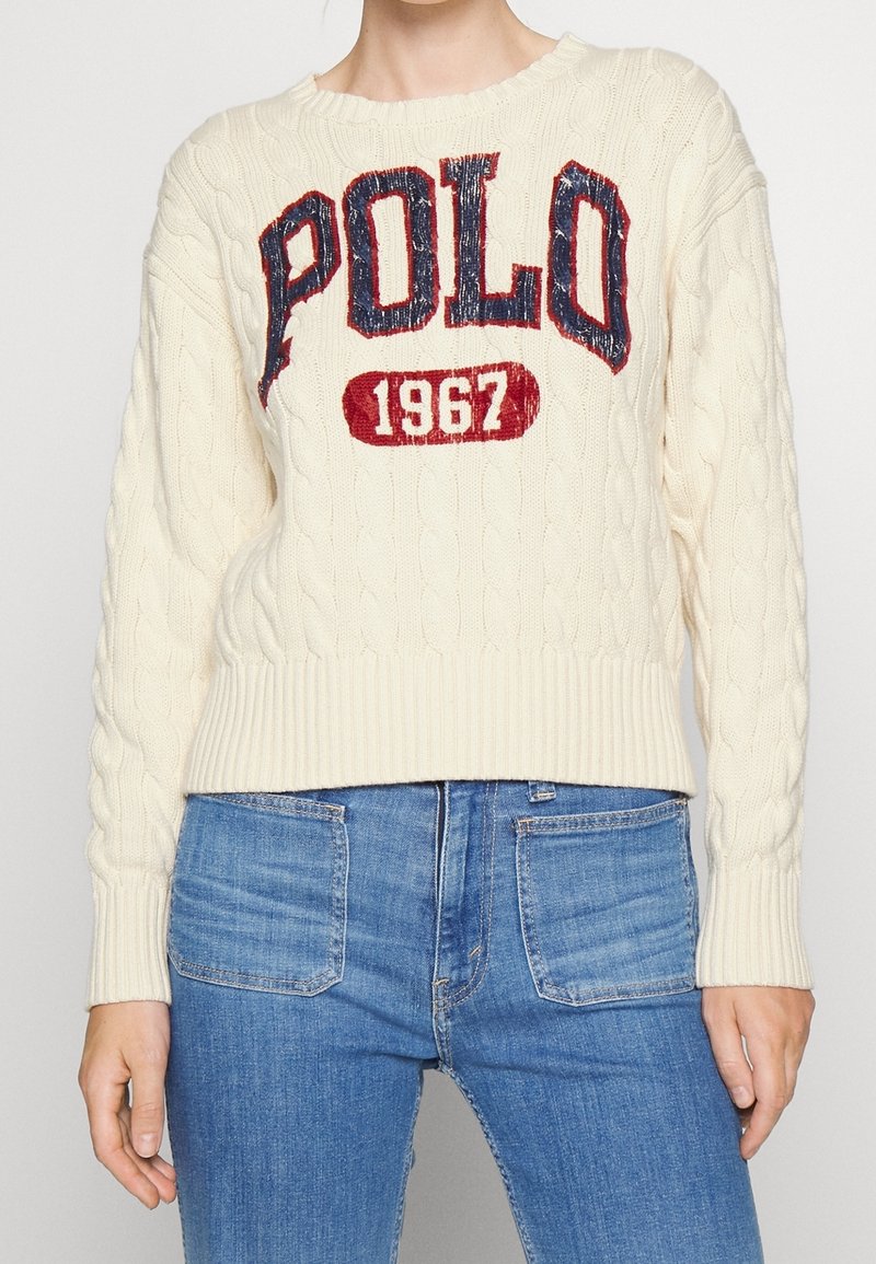 Femme portant un pull en maille torsadée de couleur crème avec l'inscription "POLO 1967" en grandes lettres rouges et bleues, associée à un jean en denim bleu.