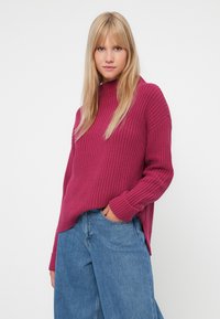 Sweater de malha em fúcsia com um decote alto, textura canelada e mangas longas, combinado com jeans de perna larga em azul claro.