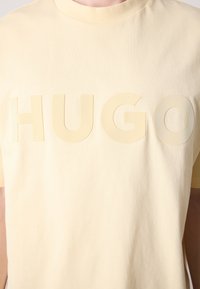 Šviesiai geltona marškinėlių su trumpomis rankovėmis pagaminti iš minkštos medvilnės. Yra pernelyg didelis "HUGO" logotipas toniniu raštu. Apvalus kaklo iškirpimas.