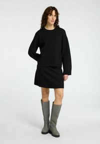 Crni pleteni pulover s malim ramenima, u kombinaciji s crnom suknjom u stilu skater. Model nosi sivo gumene čizme do koljena. Neutralna pozadina.