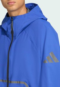 Modr�á nepromokavá bunda s kapucňou, s predným zipsom, sivými akcentmi a reflexným logom na rukáve. Hladká textúra.