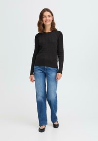 Svart ribbestrikket genser med lange ermer, kombinert med blå straight-leg jeans og svarte ballettsko, mot en enkel hvit bakgrunn.