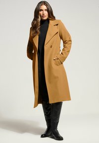Kamel-farvet uldfrakke med dobbeltradet design, brede revers, sidelommer og knælangt snit, båret over en sort sweater og bukser.