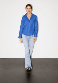 Blaue Leinenhemd mit Kragen, getragen über hellblauen Schlagjeans und schwarzen Plateauschuhen. Modell steht auf grauem Boden.