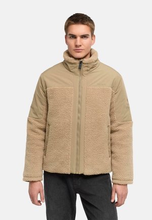 Giovane uomo che indossa una giacca in pile beige con colletto alto e pantaloni neri, in piedi davanti a uno sfondo chiaro e uniforme.