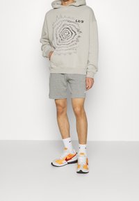 Světlá šedá mikina s grafickým spirálovým vzorem, kombinovaná se šedými šortkami a vícebarevnými teniskami. Obsahuje bílé kotníkové ponožky a má volný střih.