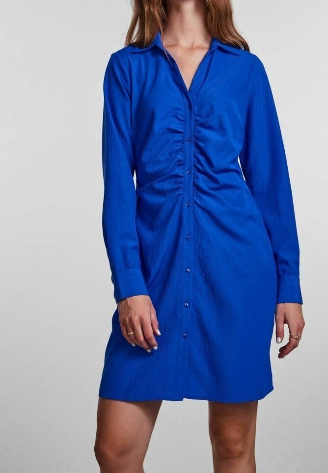 Femme portant une robe bleu roi à manches longues, boutonnée sur le devant, arrivant au genou, avec un détail froncé à la taille.