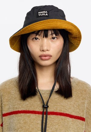 Joven con cabello oscuro lacio que lleva puesto un sombrero de cubo de pana negro y mostaza con correas para la barbilla y un suéter beige con una franja roja.