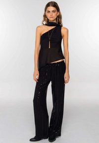 Vrouw in een een-schouder zwarte top met een lange sjaal-detail en zwarte wijde broek met verticale paillettenstrepen.