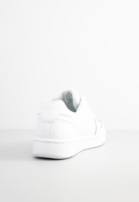 Bílé tenisky s hladkým koženým svrškem, polstrovaným límcem a jemným tonálním prošíváním. Mají odolnou gumovou podrážku a minimalistický design.