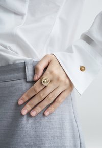 Anel dourado com design de ferradura e pedras preciosas brancas, usado numa mão contra um fundo de tecido cinzento claro texturizado.