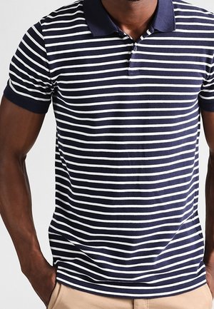 Mann trägt navyblau-weiß horizontal gestreiftes kurzärmeliges Poloshirt mit beigen Hosen, Hände in den Taschen, vor einfarbigem Hintergrund.
