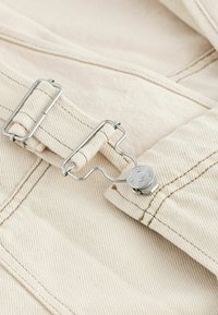 Nahaufnahme von beigem Stoff mit einer Metallniete und einem Verschluss, der an einem verstellbaren Riemen befestigt ist, wobei die Nähdetails sichtbar sind.