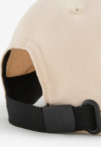 Beige vászon sapka fekete állítható pánttal, amelyen egy fekete csatt található, amelyen egy kecskebőr koponya logó van. Texturált szövetfelület.
