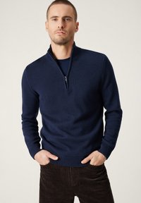 Granatowy sweter z zamkiem na szyi, z prążkowanym dołem i mankietami, wykonany z teksturowanej dzianiny. Nosi się go na dopasowaną koszulę, w zestawieniu z brązowymi spodniami ze sztruksu.