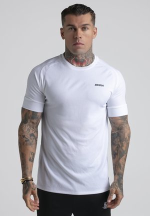 Mann med blomsterhals-tatovering og fullarmstatoveringer, iført en tettsittende hvit Siksilk-t-skjorte og svarte bukser, står mot en ensfarget bakgrunn.