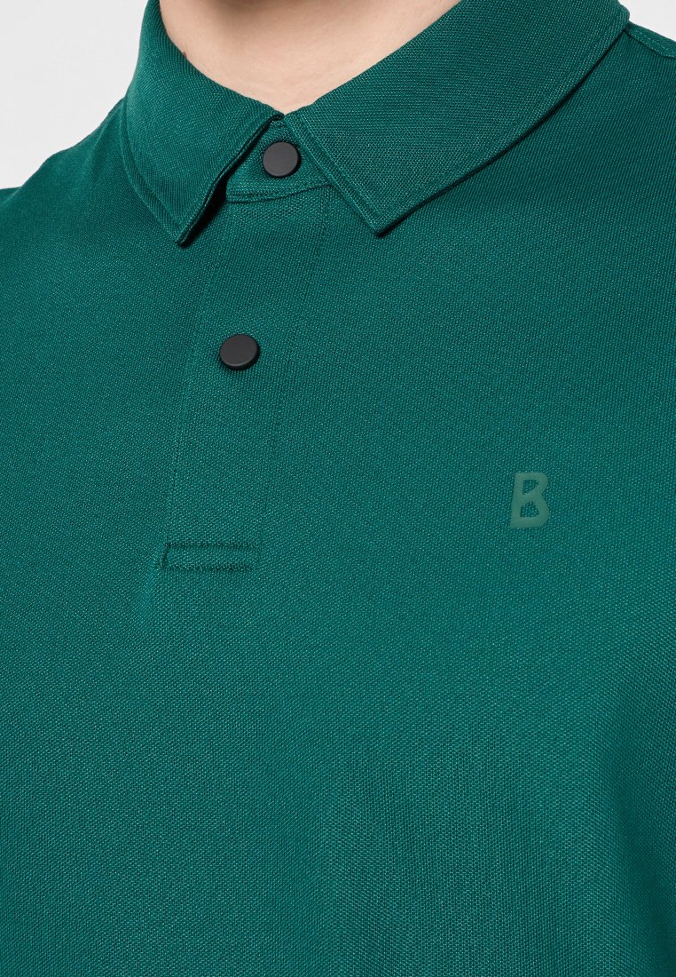 Dunkelgrünes Poloshirt aus strukturiertem Stoff, mit zwei schwarzen Knöpfen und einem dezenten Logo auf der Vorderseite. Der Kragen ist gefaltet und strukturiert.