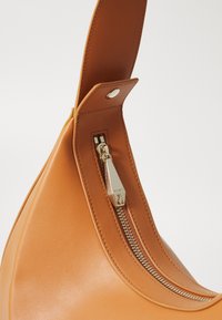 Kůže hnědé barvy s hladkým povrchem, obsahující zipové uzavírání a stříbrné kovové detaily, ukazující elegantní a zakřivený design.