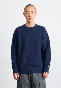 Marineblauwe gebreide sweatshirt met geribbelde ronde hals, manchetten en onderrand. Voorzien van een klein geel logo op de linker mouw. Textuurstof.