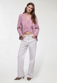 Blazer accorciato rosa chiaro con tre bottoni, abbinato a pantaloni bianchi a vita alta. La modella indossa tacchi a punta marroni con dettagli a fiocco.