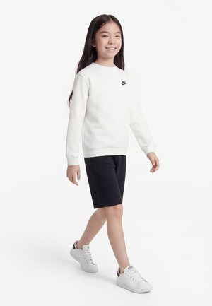 Χαμογελαστή νεαρή κοπέλα που περπατά, φοράει λευκό φούτερ Nike, μαύρα σορτς και λευκά αθλητικά παπούτσια, σε φόν από απλό λευκό τοίχο.