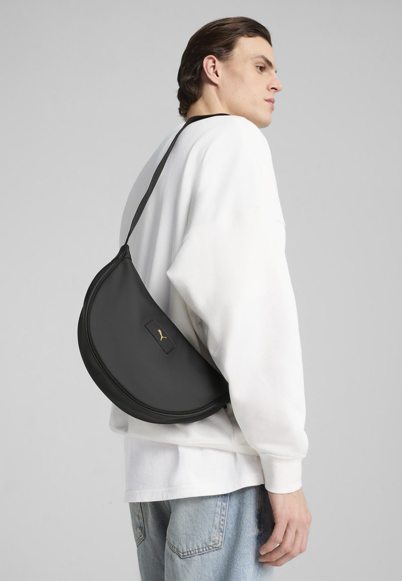 Black leather bag with a half-moon shape, featuring a small gold logo. Smooth texture and narrow shoulder strap. Worn over a white sweatshirt.