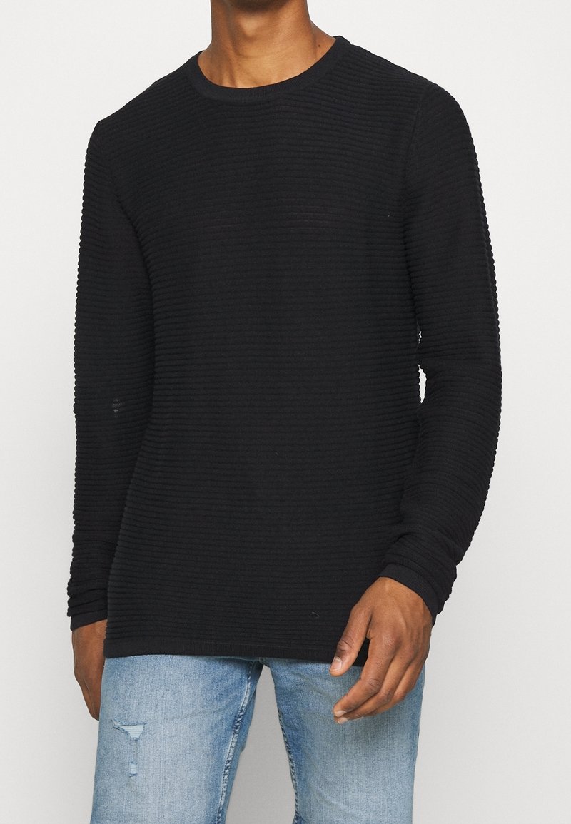 Czarny sweter z długim rękawem o fakturowanym prążkowanym wzorze, z okrągłym dekoltem i dopasowanymi mankietami, w połączeniu z jasnoniebieskimi, przetartymi dżinsami.