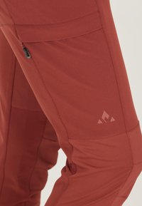 Röda utomhusbyxor i lätt tyg, med en ficka med dragkedja och en liten logotyp. Designen är figursydd med artikulerade knän.