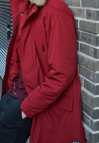 Röd puffjacka med en pälskantad huva, fickor med dragkedja och en texturerad yta, bärs över en mörk skjorta och byxor mot en tegelvägg.