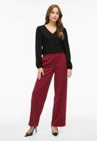 Top nero lavorato a maglia con scollatura a V e maniche a palloncino, abbinato a pantaloni larghi bordeaux e décolleté neri a punta con dettagli dorati.