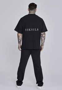 Camisa negra de gran tamaño con el logotipo blanco "SIKSILK" en la parte trasera, combinada con pantalones jogger negros y zapatillas blancas. Tejido de algodón suave.