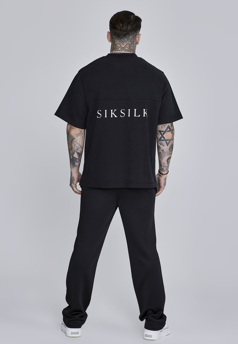 Camisa negra de gran tamaño con el logotipo blanco "SIKSILK" en la parte trasera, combinada con pantalones jogger negros y zapatillas blancas. Tejido de algodón suave.