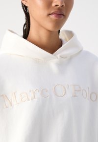 Kreem värvi kapuutsiga sviiter, millel on heledamas toonis tikitud tekst "Marc O'Polo", pehme tekstuuriga ja laia, struktureeritud kapuutsiga.