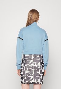 Lichtblauwe cropped trui met een hoge kraag en zwarte logo-accenten, gecombineerd met een geblokte zwart-witte minirok.