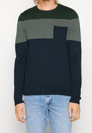 Homme portant un pull à manches longues à blocs de couleurs avec une bande grise à motifs verts, un bas marine, une poche poitrine, et un jean bleu clair.
