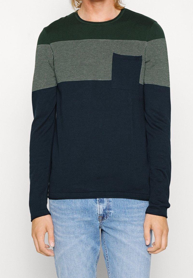 Homme portant un pull à manches longues à blocs de couleurs avec une bande grise à motifs verts, un bas marine, une poche poitrine, et un jean bleu clair.