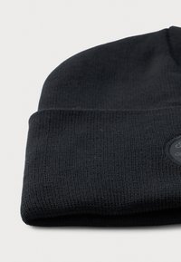 Căciulă tricotată neagră cu manșetă pliată și un patch rotund negru pe manșetă, așezată pe o suprafață albă.