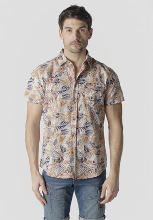 Hombre con camisa de manga corta abotonada con patrón de hojas en naranja, beige y azul marino, y pantalones cortos de mezclilla azul, de pie mirando hacia adelante.