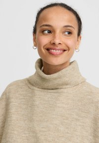 Beige coltrui met een zachte, geribde kraag. De stof heeft een subtiele textuur, met een ontspannen pasvorm en een neutrale tint voor het laagjeslook.