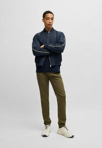 Navy bomber jacket with contrasting white stripes, paired with olive green pants and light-colored sneakers. Smooth material, casual fit.