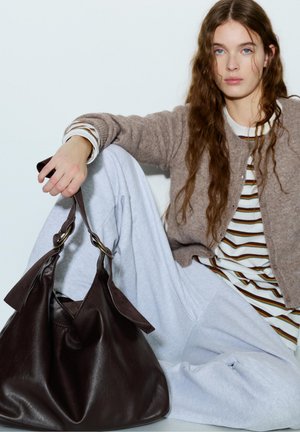 Jonge vrouw met lang golvend haar zit en houdt een donkerbruine leren handtas vast, draagt een beige cardigan, gestreept shirt en lichtgrijze broek tegen een witte achtergrond.