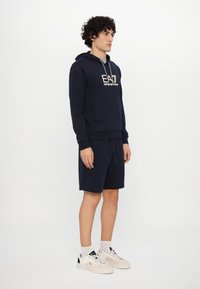 Navy hoodie with front pocket and logo, paired with matching shorts. Model wears white sneakers with black accents. Simple and sporty design.