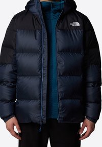 Veste matelassée bleu marine et noire avec capuche, dotée d'une fermeture éclair et d'une doublure intérieure bleue à pois. Comprend des poignets élastiques et un détail logo.