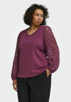 Kvinde med krøllet hår bærer en lilla langærmet top med blondeærmer og sorte bukser, poserer med den ene hånd i lommen.