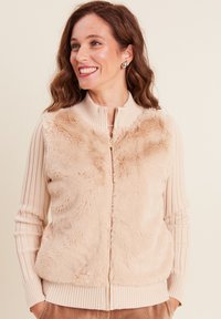 Gilet zippé beige avec un devant en fausse fourrure douce, des manches en tricot côtelé et un col haut. Il possède une fermeture éclair dorée et une texture lisse.