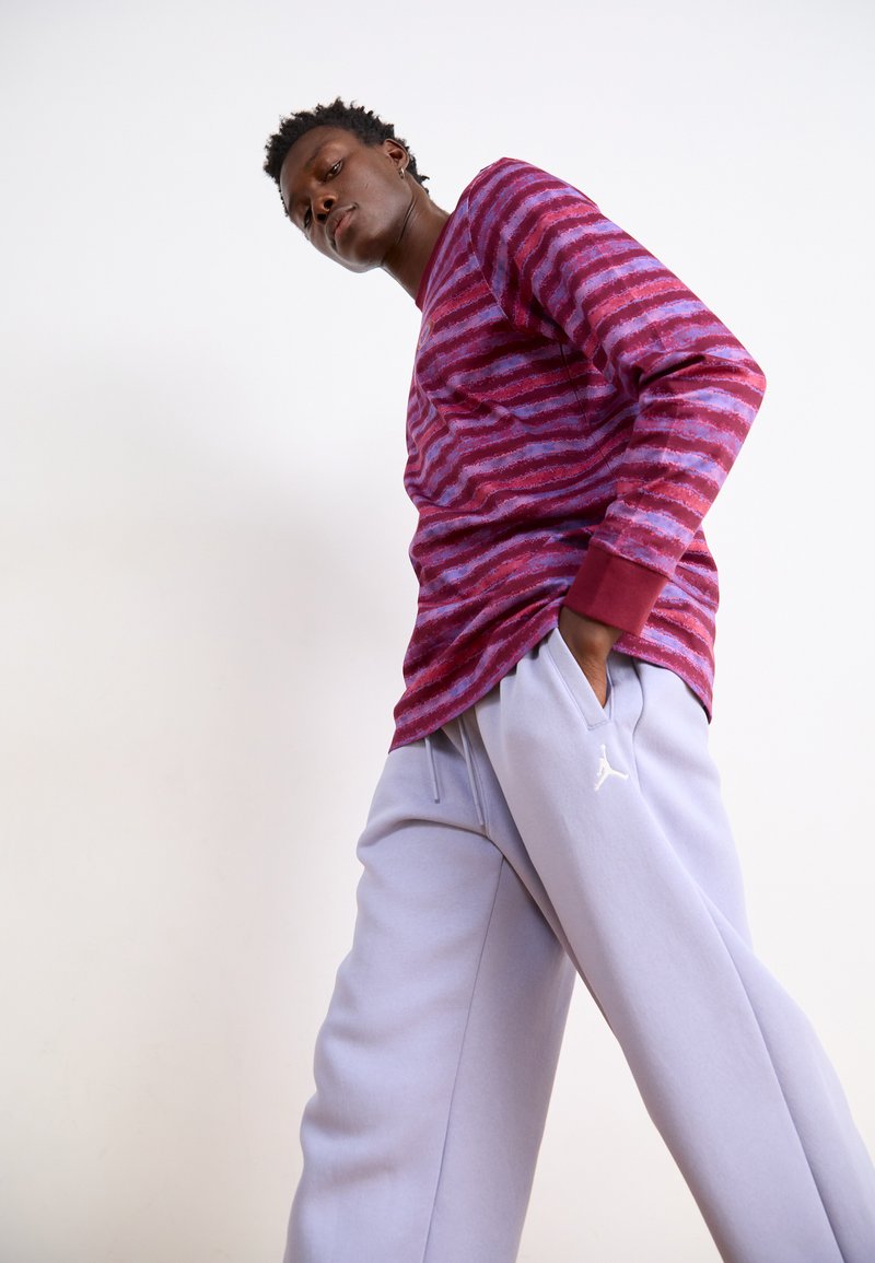 Chemise à manches longues rayée rouge et violette, associée à un pantalon de jogging gris clair arborant un logo sur la jambe gauche. Le tissu semble doux et texturé.