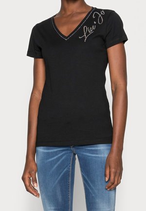 Femme portant un t-shirt noir à col en V avec un texte en strass sur l'épaule gauche et un jean bleu délavé, sur fond uni.