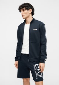 Námornícka modrá bunda na zips so zvislými pruhmi, bielym logom a zodpovedajúce šortky s grafickou potlačou. Mäkký, ľahký materiál.