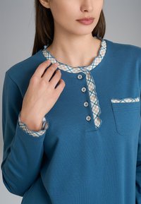 Maglietta a maniche lunghe blu con colletto a onde, caratterizzata da un motivo a quadri all'interno e una tasca a toppa. Cinque bottoni decorano la parte anteriore.